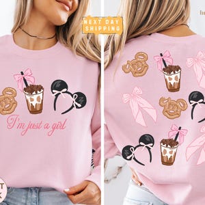 May include: Pale pink sweatshirt featuring cartoon illustrations of a pretzel, a drink, and a Minnie Mouse ear headband. The front of the shirt has the text "I'm just a girl". The back of the shirt has the same graphics.