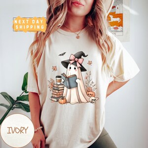 May include: An ivory short-sleeve t-shirt featuring a graphic of a ghost wearing a witch hat with a pink bow, reading a book. The design includes a stack of books, a coffee cup, pumpkins, a skull, autumn leaves, and a bat.
