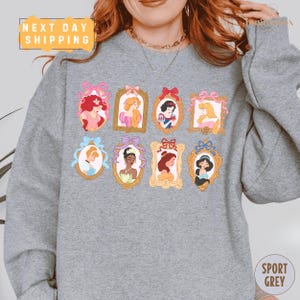 A person wearing a gray sweatshirt with a collage of illustrations of Disney princesses arranged in a grid pattern.