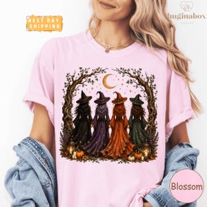 A woman wearing a pink t-shirt with a graphic design depicting four women dressed in witch costumes standing in a circle under a crescent moon.