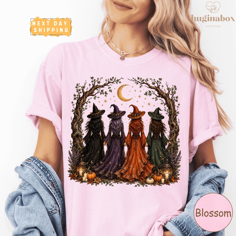 A woman wearing a pink t-shirt with a graphic design depicting four women dressed in witch costumes standing in a circle under a crescent moon.