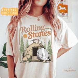 May include: Cream-colored t-shirt featuring the text "Rolling Stones" in a retro font, with an illustration of a tomb and three crosses. The design includes a sun, pine trees, and the text "Since 4D 33".