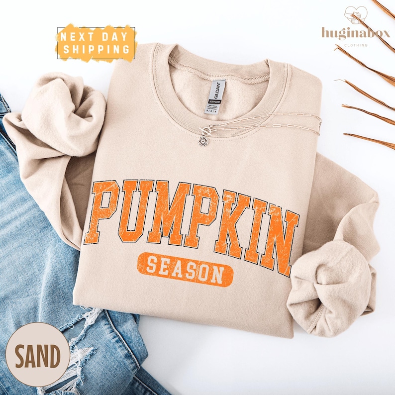 a beige sweatshirt with the word "PUMPKIN" printed in orange letters, along with the word "SEASON" below it. The sweatshirt is laid out on a white background, with a pair of blue jeans visible in the background. There is also a small text box in the top left corner that reads "NEXT DAY SHIPPING".