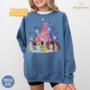 A woman wearing a blue sweatshirt with a colorful illustration of Disney princesses and a castle on it.