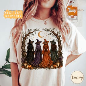 A woman wearing a white t-shirt with a graphic design depicting four women in witch costumes standing under a crescent moon and surrounded by autumn foliage.