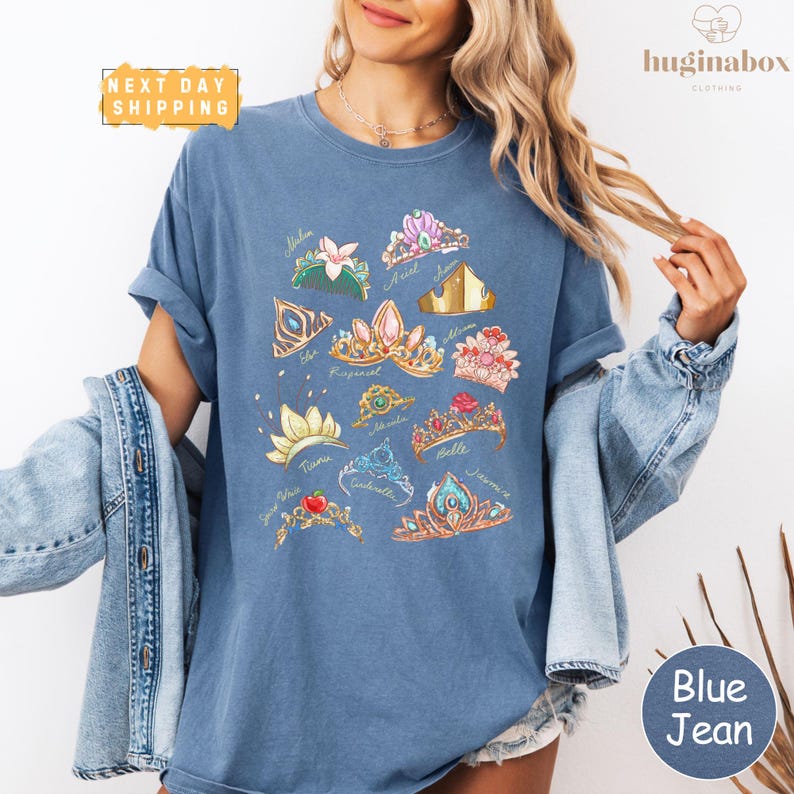 A woman wearing a blue t-shirt with a graphic design of various crowns and jewelry, paired with denim jeans and a denim jacket.