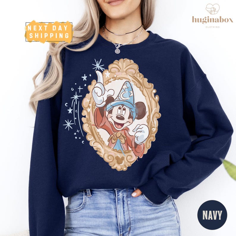 A woman wearing a navy blue sweatshirt with a Mickey Mouse design and a gold frame, standing against a white background.