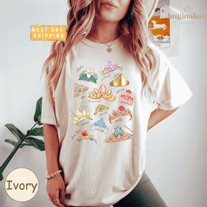 A woman wearing a white t-shirt with a colorful graphic design of various crowns and jewels printed on it.