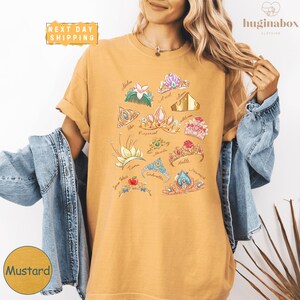 A woman wearing a yellow t-shirt with a graphic design of various crowns and floral elements. She is also wearing a denim jacket and has her hair styled in loose waves.