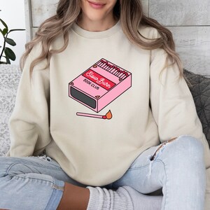 May include: A light beige sweatshirt with a graphic of a pink matchbox with the text "Slow Burn Book Club" and a lit match below it.