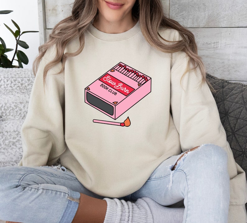 May include: A light beige sweatshirt with a graphic of a pink matchbox with the text "Slow Burn Book Club" and a lit match below it.