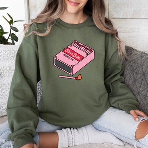 May include: A green sweatshirt with a pink box of matches that says "Slow Burn Book Club" and a lit match on the front.