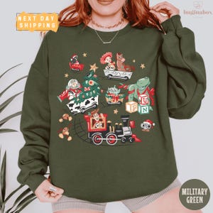 May include: Military green sweatshirt featuring a festive graphic of Toy Story characters on a train and in various holiday-themed vehicles. The design includes a Christmas tree, stars, and the text "FUN" on blocks. The words "NEXT DAY SHIPPING" are also visible.