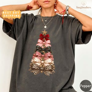 May include: A charcoal grey short-sleeved t-shirt featuring a graphic of a Christmas tree constructed from various patterned bows, including red, plaid, striped, polka-dot, and leopard print designs, topped with a gold star.