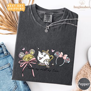 a gray t-shirt with a Disney-themed design, including a teapot, a character, and various other elements. The t-shirt is displayed on a white background, along with a pair of jeans and a necklace.