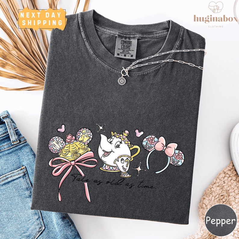 a gray t-shirt with a Disney-themed design, including a teapot, a character, and various other elements. The t-shirt is displayed on a white background, along with a pair of jeans and a necklace.