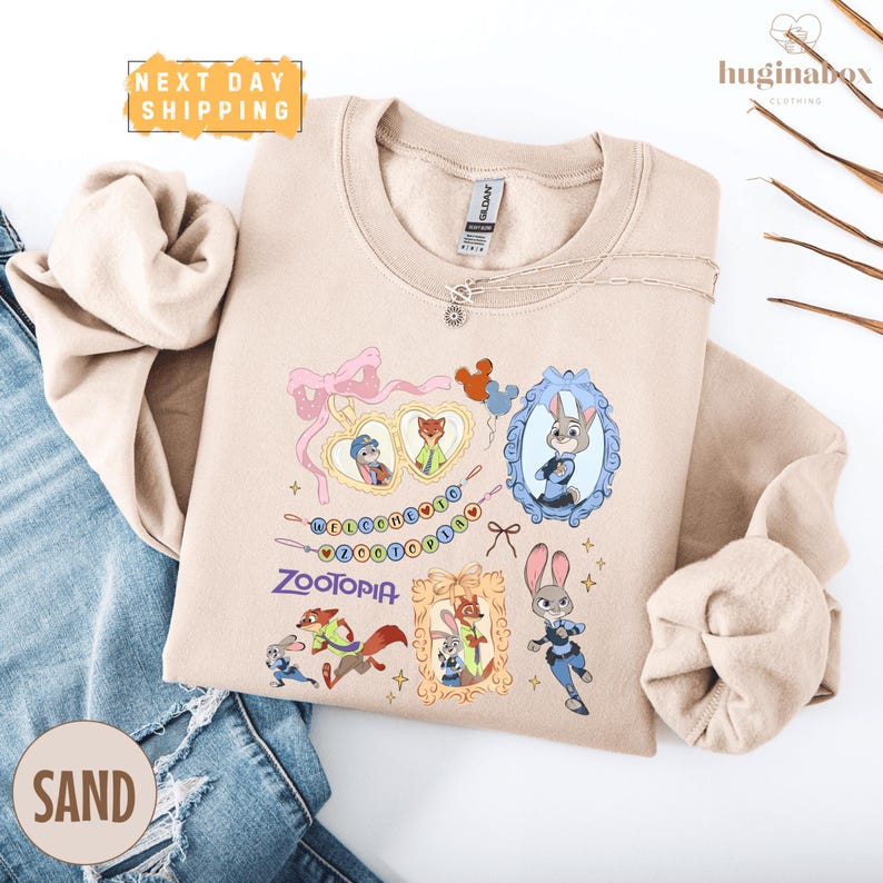 A beige sweatshirt with a cartoon character design, including a rabbit, a fox, and other animals, along with a heart and the word "Zootopia". The sweatshirt is displayed on a white background, and there is a pair of blue jeans visible in the image.