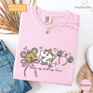 a pink t-shirt with a cartoon character design, a pink ribbon, and the text "Tale as old as time." The t-shirt is displayed on a white background, along with a pair of jeans and some accessories.
