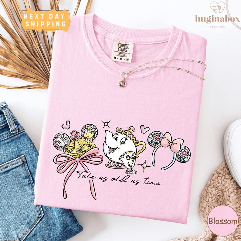 a pink t-shirt with a cartoon character design, a pink ribbon, and the text "Tale as old as time." The t-shirt is displayed on a white background, along with a pair of jeans and some accessories.
