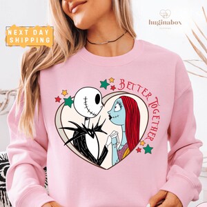 A woman wearing a pink sweatshirt with a graphic design featuring the characters Jack Skellington and Sally from the movie "Jack Skellington and Sally".