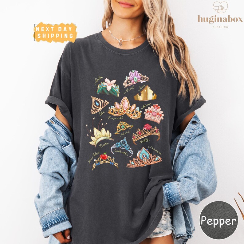 A woman wearing a black t-shirt with a graphic design of various crowns and jewelry, paired with a denim jacket and a necklace.