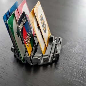 May include: A close-up of a UNO card holder with a variety of cards, including a wild card, a draw four card, and several number cards. The holder is made of gray plastic and has a slot for each card.