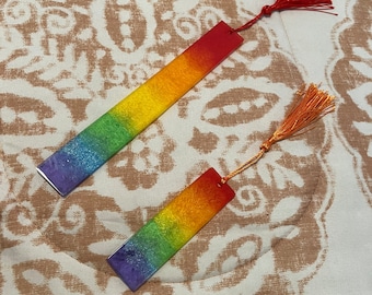 May include: Two rainbow glitter bookmarks with tassels. The bookmark on the left has a red tassel and the bookmark on the right has an orange tassel.