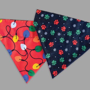 May include: Reversible dog and cat bandana with Christmas lights and paw prints. One side features a red background with colorful Christmas lights. The other side has a navy blue background with green and red paw prints and white snowflakes. The text "Christmas Lights Reversible Dog + Cat Bandana" is at the top.