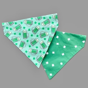 May include: Two green dog bandanas. One bandana has a pattern of green beer mugs and shamrocks on a light green background. The other bandana has a pattern of white shamrocks on a dark green background.