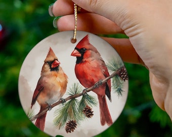 May include: A round ceramic ornament featuring a watercolor painting of two red cardinals perched on a pine branch with pine cones. The cardinals are painted in shades of red, brown, and white.