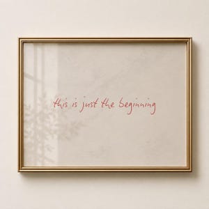 May include: A rectangular wall art piece featuring a thin gold frame surrounding a light beige background. The text "this is just the beginning" is written in the centre in a handwritten red script font, with a subtle shadow of foliage.