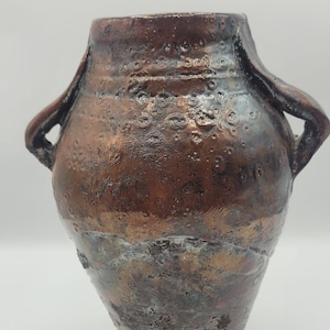 Puede incluir: Un jarrón de cerámica rústico de color cobre con dos asas. El jarrón tiene una superficie texturizada y un acabado desgastado.