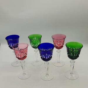 May include: Set of seven colorful crystal wine glasses. Each glass features a different colored bowl, including blue, pink, and green. The glasses have a clear stem and base, and are arranged on a white surface.