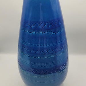 May include: A tall, tapered ceramic vase in a vibrant shade of blue with a glossy finish. The surface features horizontal bands of embossed geometric patterns, including diamonds, circles, and dotted lines, creating a textured, repeating design.