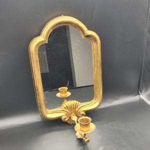 May include: A gold-colored wall mirror with a scalloped top and a decorative candle holder. The mirror has a rectangular shape with rounded edges. The candle holder features two candle cups and a seashell design.