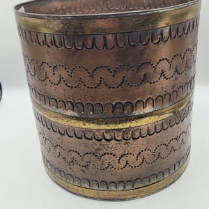 May include: A copper-colored metal container with a decorative design. The container has a rounded oval shape with a brass-colored band at the top and bottom. The body features a repeating pattern of curved lines and dotted details.