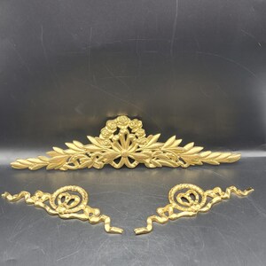 May include: Three gold-colored decorative architectural elements. The largest piece features a floral and leaf design. Two smaller, matching pieces have a scroll and swirl design. All pieces are made of a shiny, metallic material.