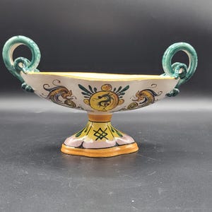 May include: A decorative ceramic bowl with a pedestal base and two curved handles. The bowl is painted with a yellow, green, and orange color scheme, featuring dragon and floral designs. The base is approximately 4 inches wide.