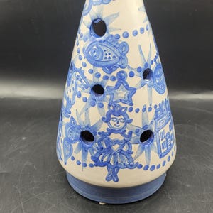May include: A conical ceramic sculpture with a white base and blue hand-painted illustrations of a figure, fish, stars, and geometric patterns. The object features several circular cutouts distributed across its surface and a solid blue base.