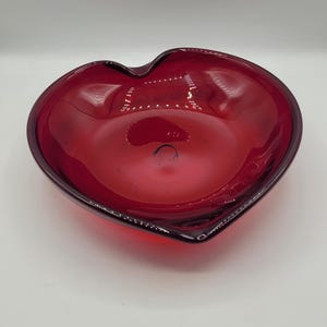May include: A heart-shaped bowl crafted from translucent red glass. The bowl features smooth, curved edges and a deep, rounded basin, with a glossy finish that reflects light across its vibrant, solid red surface and uniform color throughout.