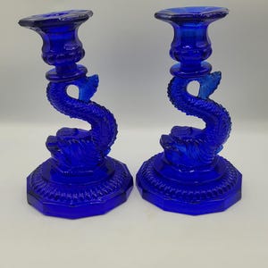 May include: A pair of cobalt blue glass candle holders. Each has a dolphin stem and an octagonal base. The candle holders are approximately 20 cm tall. A decorative item for the home.