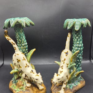 May include: A pair of ceramic candle holders featuring cream-colored leopards with brown spots. Each leopard is positioned at the base of a green textured palm tree. The base of each holder is brown and textured, resembling rocky ground.
