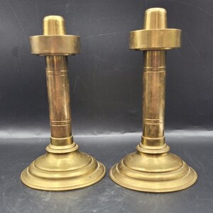 May include: Two antique brass candlesticks. Each candlestick has a round base, a cylindrical stem, and a top with a circular platform. The candlesticks are made of brass and have a warm, golden color. They are designed to hold candles.