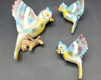 May include: Three ceramic bird figurines in shades of blue, yellow, and pink. The birds have outstretched wings and are perched on a brown branch. The figurines are on a dark gray surface.