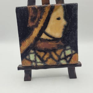 May include: A square ceramic tile featuring a profile of a person with a head covering, set on a small wooden easel. The tile has a dark background with brown, orange, and green accents. The easel is made of dark wood.