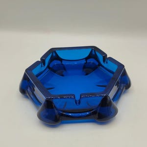 May include: A hexagonal, cobalt blue glass ashtray. The ashtray has a deep bowl with four curved sides and a flat rim with four cigarette rests. The ashtray is a vintage piece, likely from the mid-20th century.