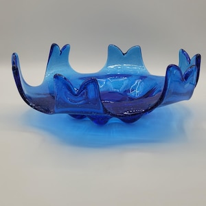 May include: A vibrant blue glass bowl with a unique, undulating rim. The bowl's design features a series of upward curves, creating a wave-like appearance. The glass has a glossy finish, and the bowl is set against a plain background.