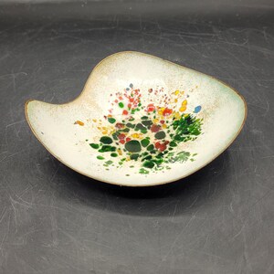 May include: A decorative, off-white enamel dish with a unique, asymmetrical shape. The dish features a colorful floral design with red, green, yellow, and blue accents. The rim is trimmed with a thin, bronze-colored metal.