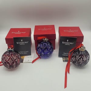 May include: Three Waterford crystal Christmas ornaments in red and blue hues, each with a silver cap and red ribbon. The ornaments are displayed with their original red boxes and informational cards.