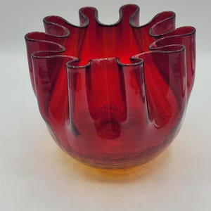 Może przedstawiać: Żywy, czerwony szklany wazon z falistym brzegiem. Wazon ma unikalny, organiczny kształt z teksturowaną powierzchnią. Krawędź jest obrysowana na czarno, dodając kontrastujący detal. Wazon jest prawdopodobnie vintage.
